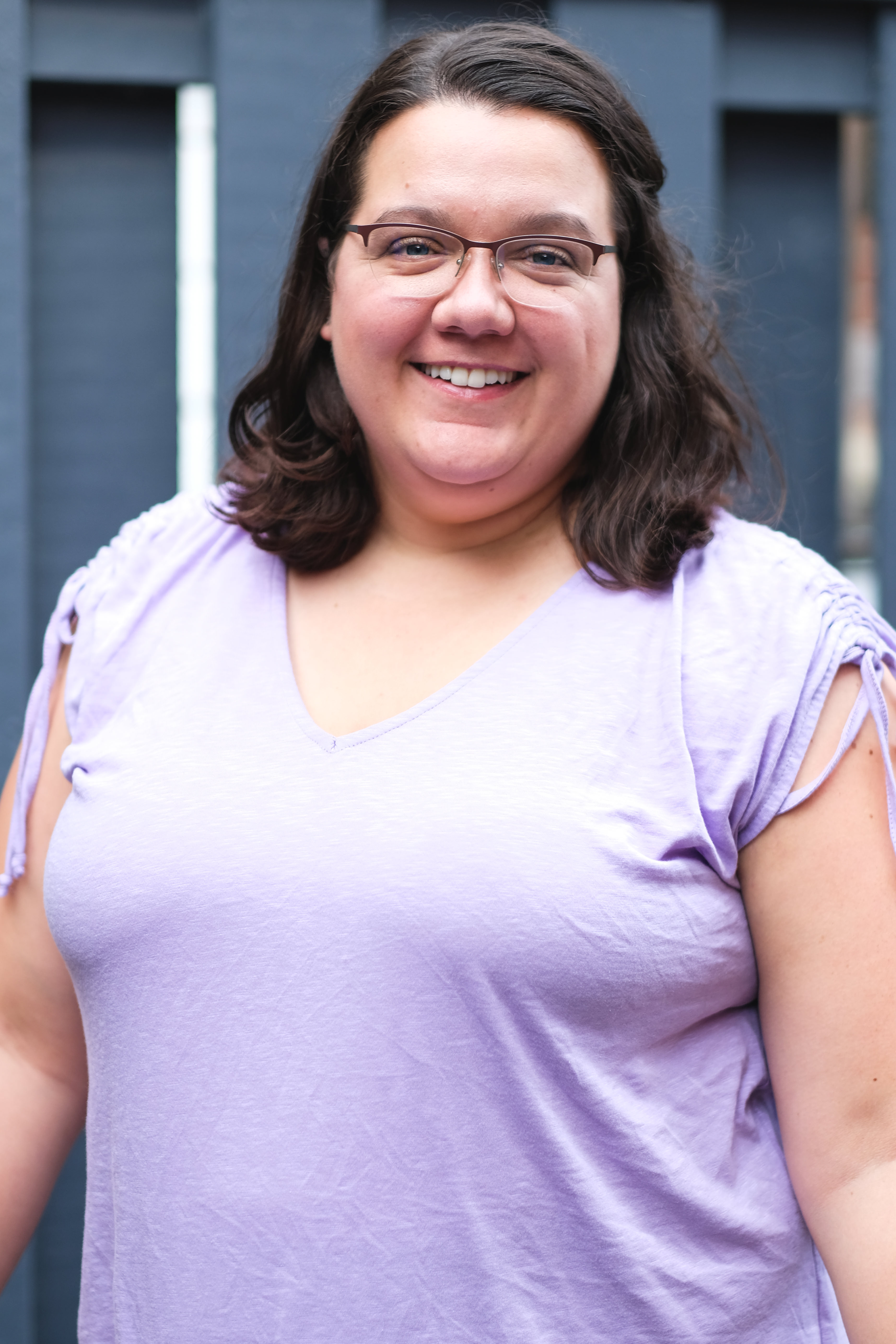 Headshot of Lisette, smiling and looking at camera. She is a white woman with medium length brown hair. She is wearing a purple short sleeved shirt.