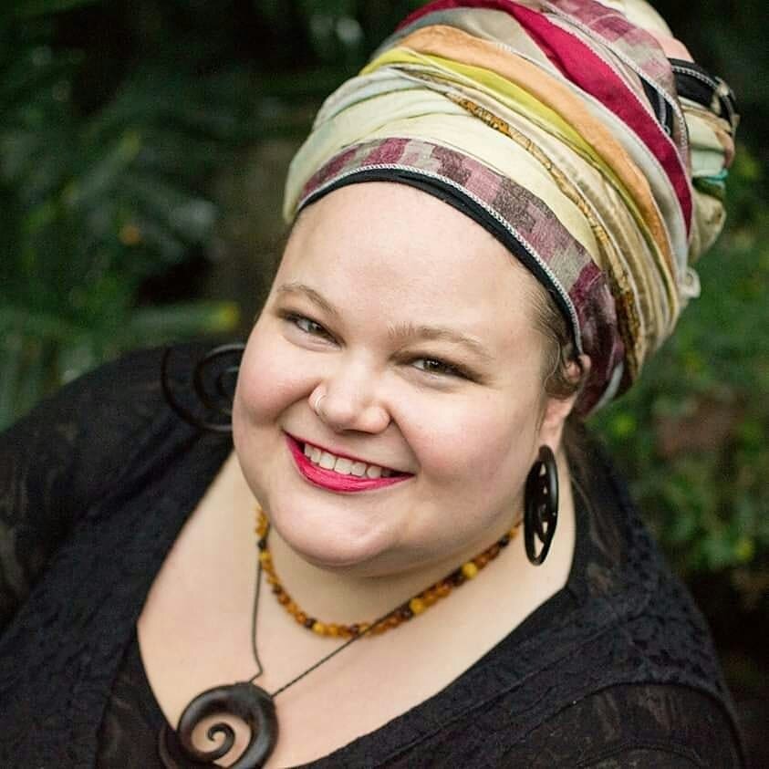 There is a photo of Beth who is a white woman smiling at the camera. She wears a colourful head scarf, a black shirt, curled earrings, and two necklaces.