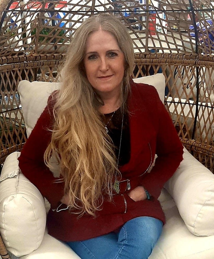Anna is a white woman with long blond hair, she is sitting in a wicker egg chair wearing a red blazer and jeans.