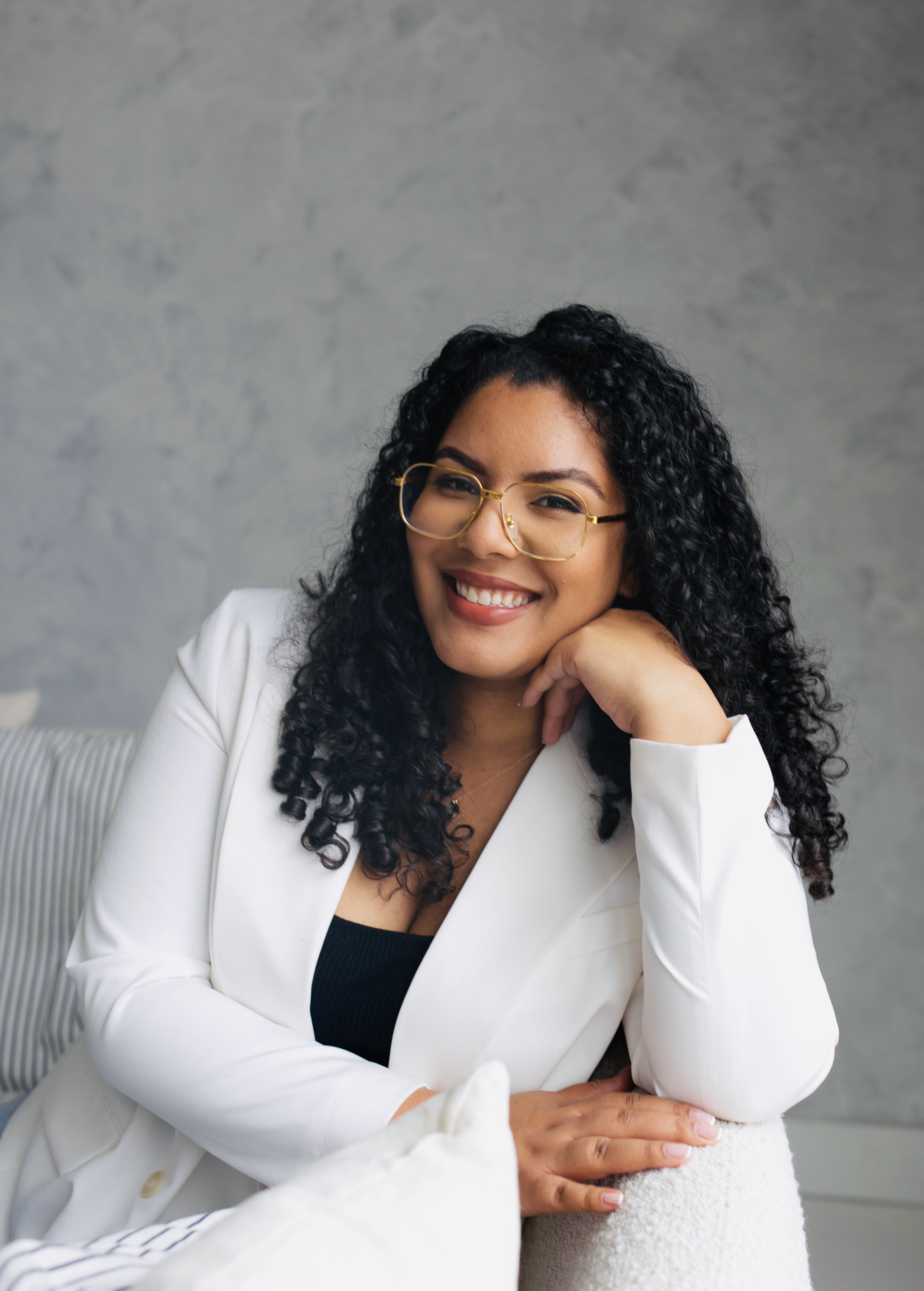 Cheyenne is a black woman with black curly hair and glasses. She is sitting on a white couch, wearing a white suit and black shirt while smiling at the camera.