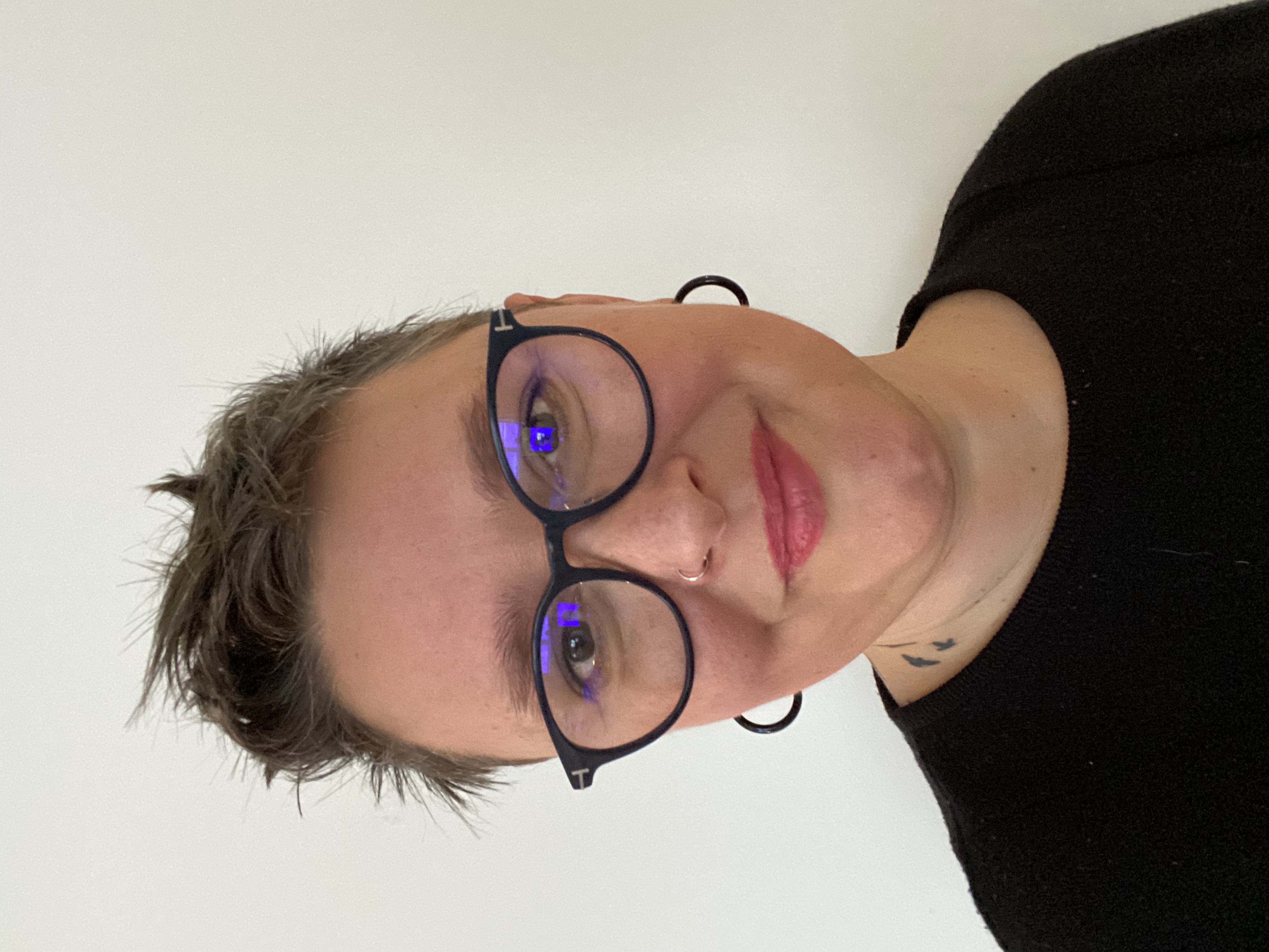 Annette is a woman woman with short hair and glasses. She is wearing a black shirt with a white wall behind her.