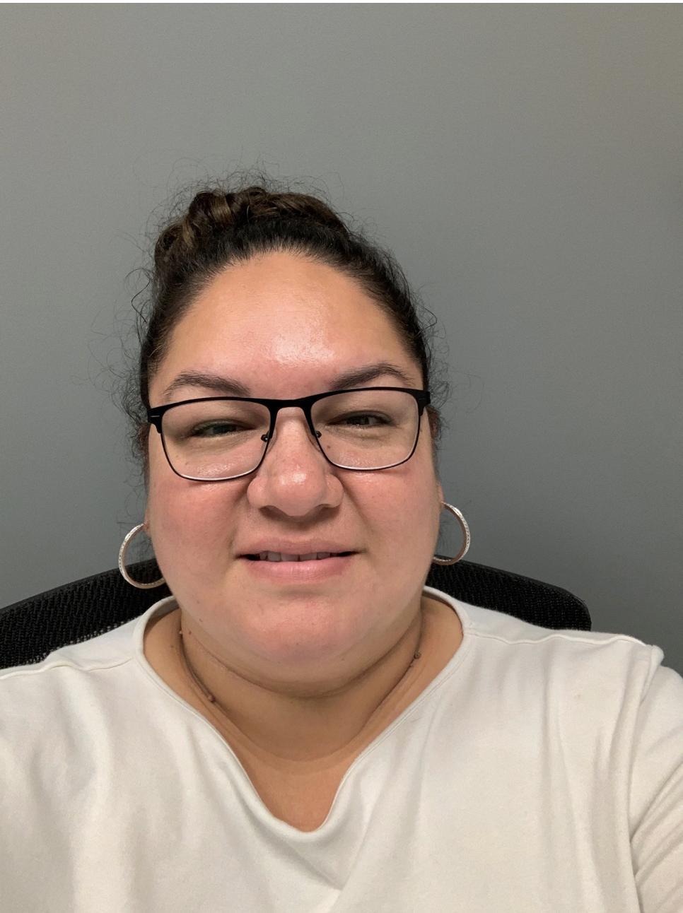 Trista Hill is an Indigenous woman with dark hair in a bun, wearing glasses and large hoop earrings. She is wearing a white shirt. You can see the top of her chair and a grey wall behind her.