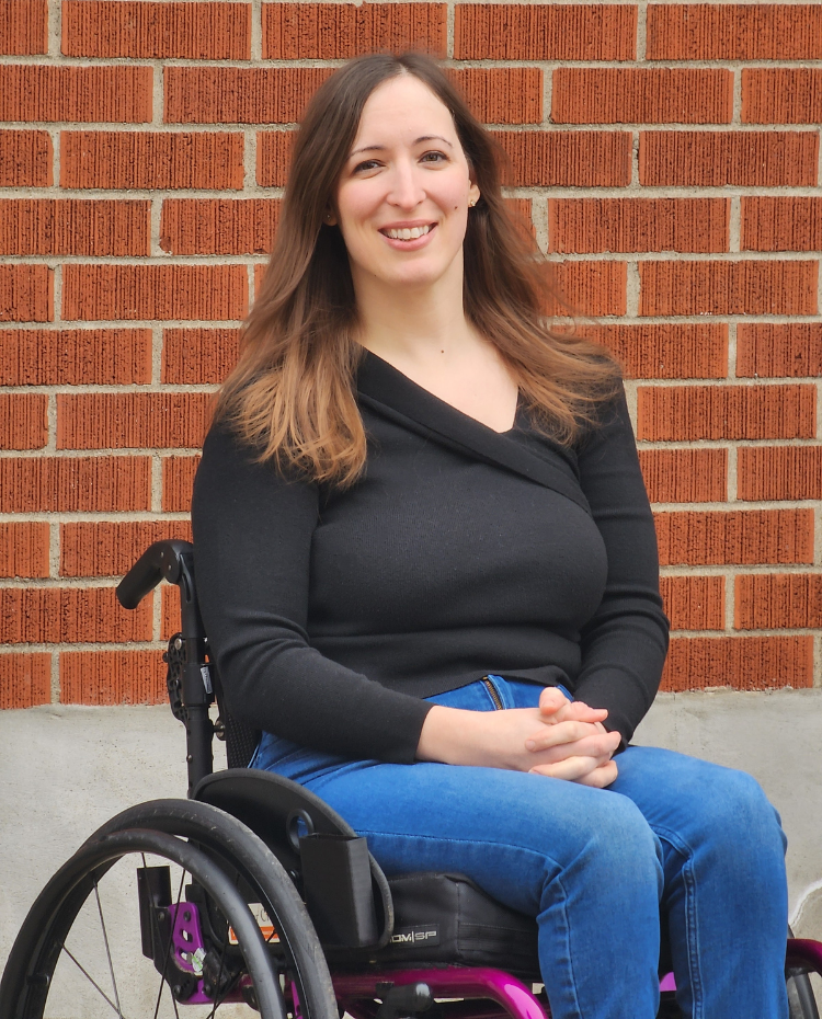 Leanne is a white woman with brown hair. She is wearing a black shirt and jeans while sitting in a wheelchair in front of a brick wall.