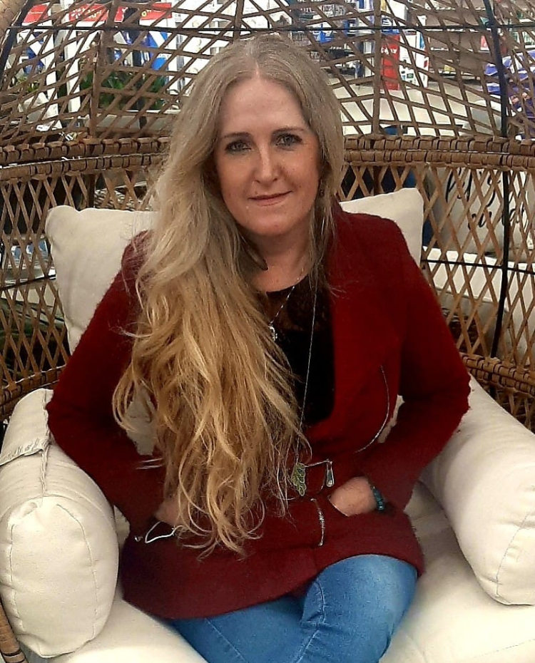 Anna is a white woman with long blonde hair. Her hands are tucked into her red jacket while she sits in a wicker chair.