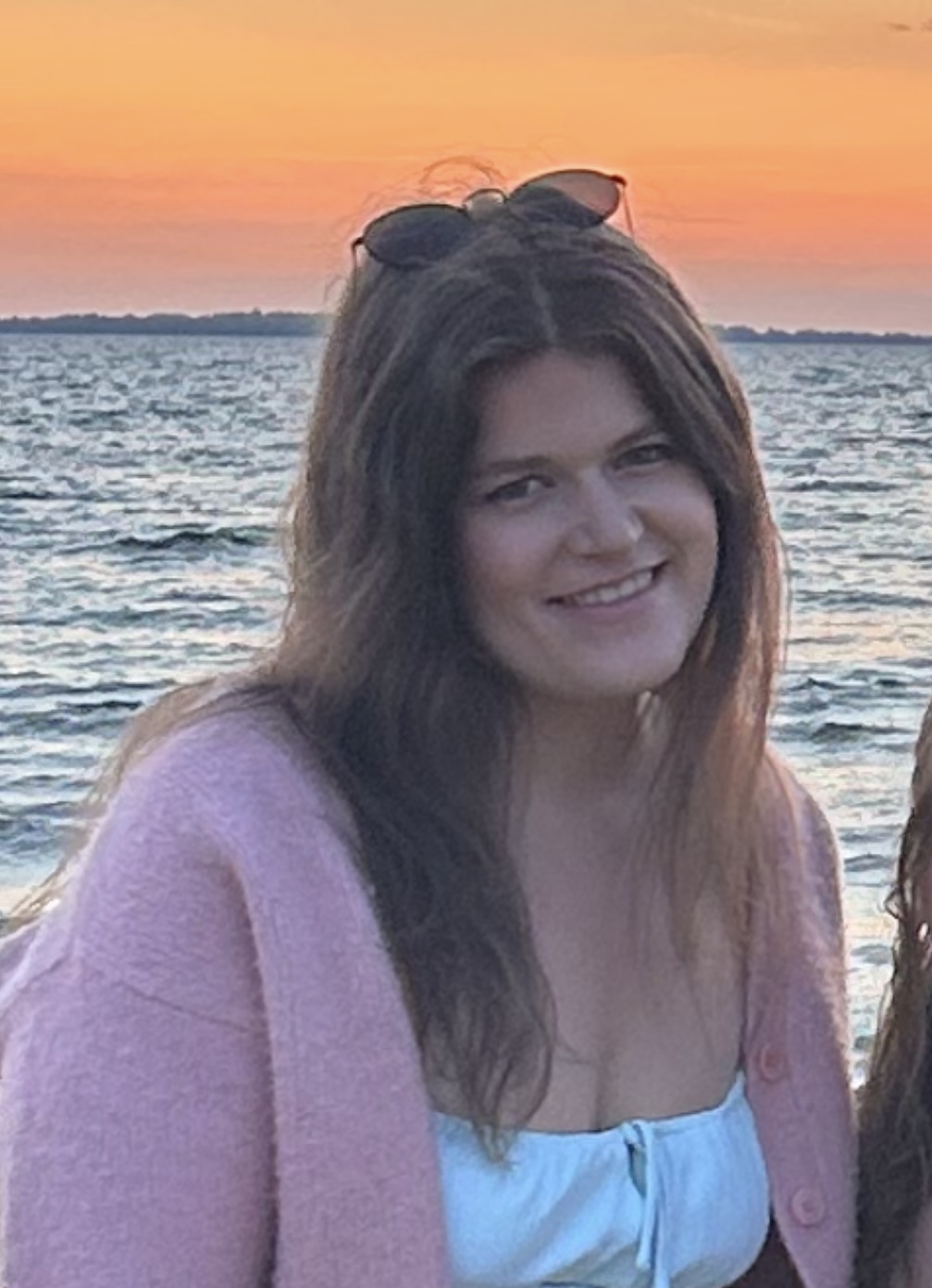 Nicole is a white woman with long brown hair. She is wearing a pink shirt and sunglasses in her head. She' standing in front of a lake at sunset.