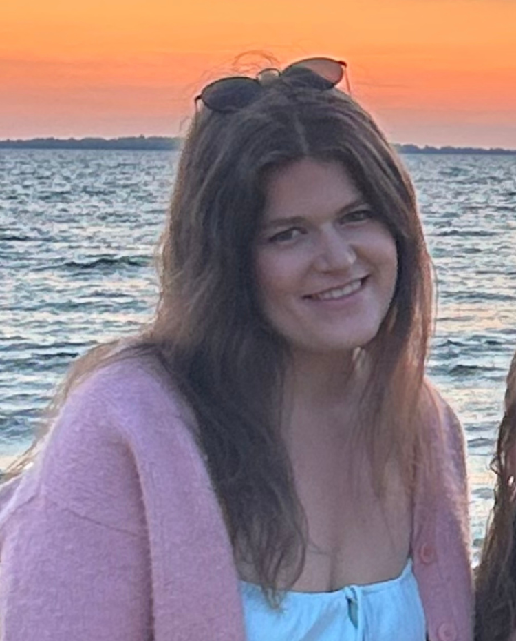 Nicole is a white woman with long brown hair who is wearing a pink sweater over a white shirt. She is standing in front of a lake at sunset with sunglasses on her head.