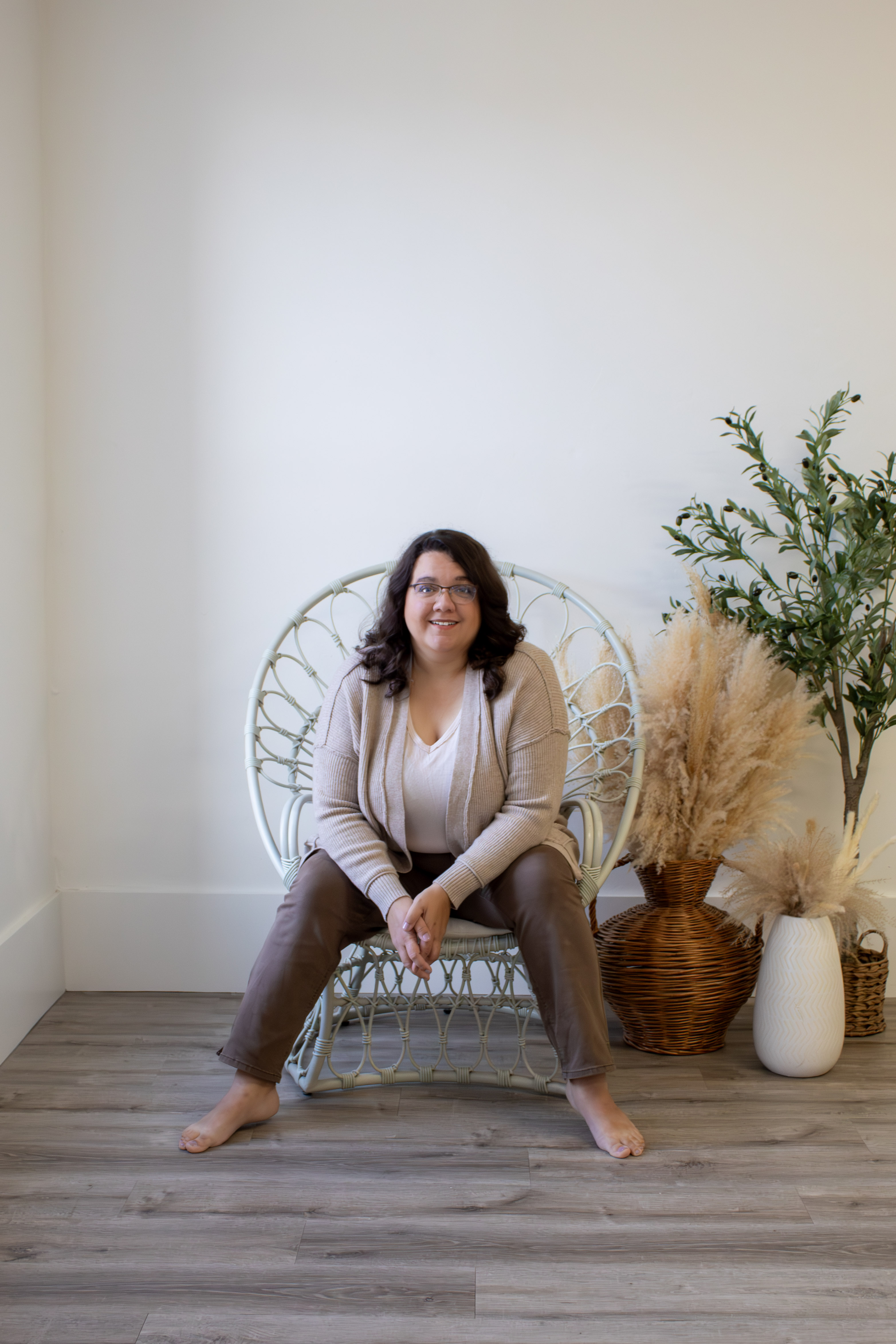 Lisette is sitting on a white wicker chair. She is wearing a beige sweater over a white shirt and brown pants. 